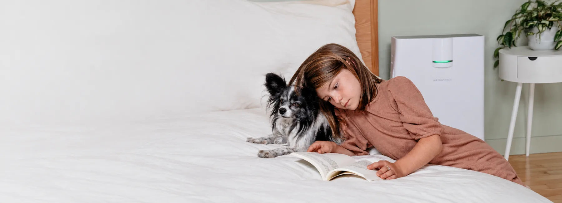 A child reading a book on a bed with a small dog, beside a Hathaspace air purifier